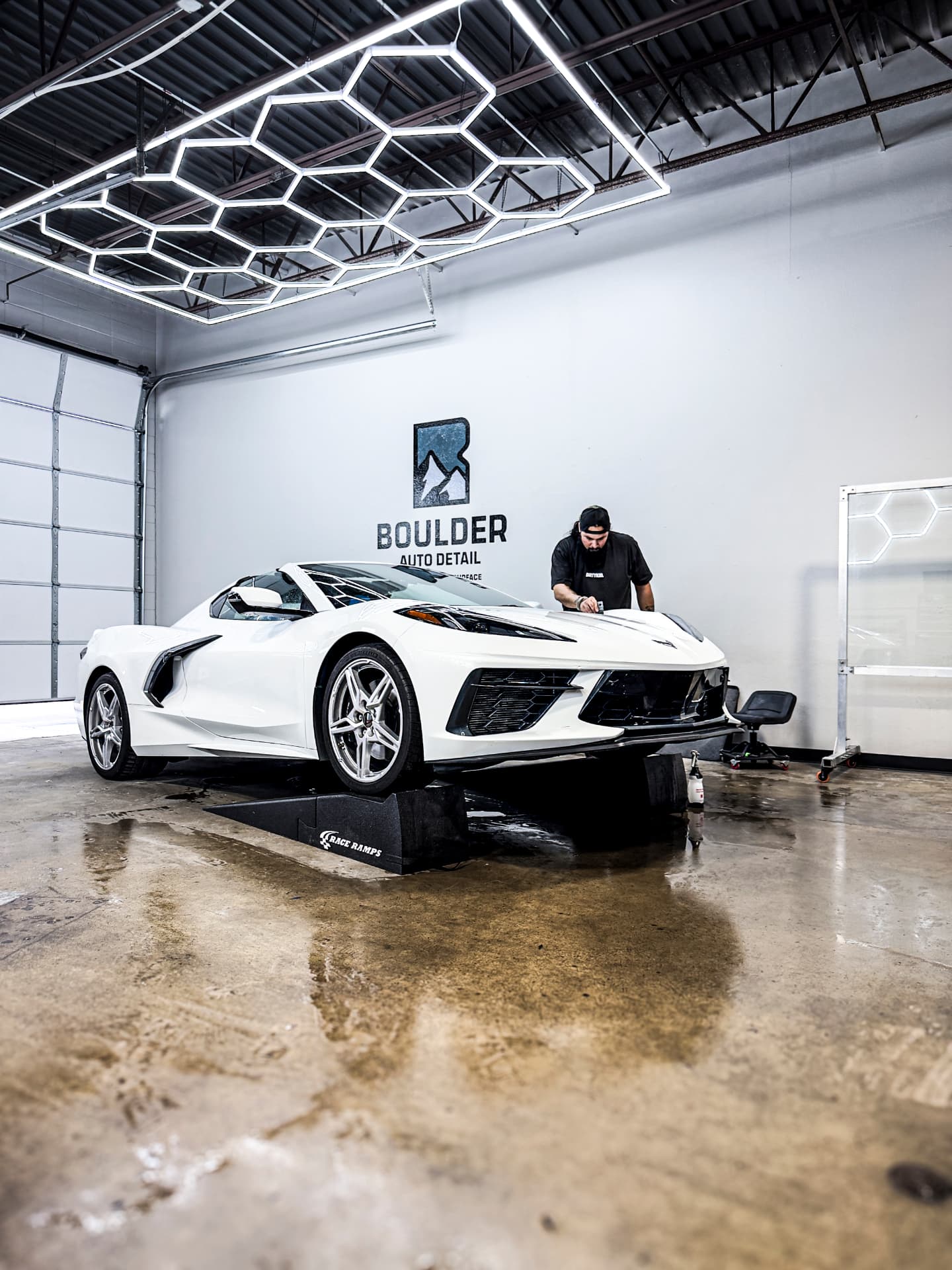 Technician protecting the hood of a white Chevrolet Corvette raised on ramps inside the Boulder Auto Detail workshop
