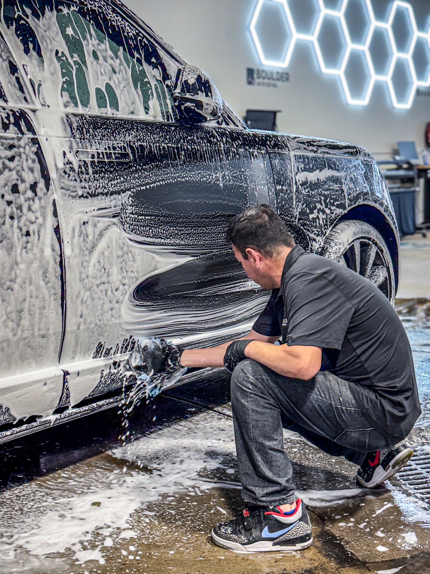Detailer hand-washing the foam-covered lower panel of a dark SUV inside the Boulder Auto Detail workshop